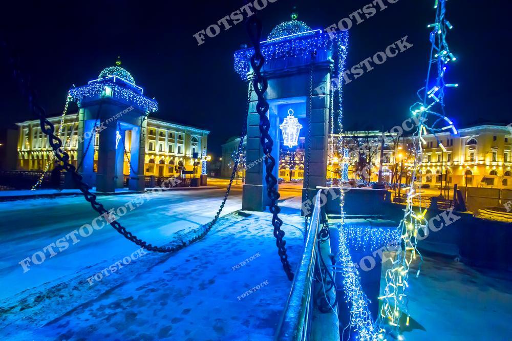 На фотографии запечатлен мост Ломоносова в Санкт-Петербурге, украшенный новогодними огнями. Гирлянды и подсветка создают волшебную атмосферу, подчеркивая детали архитектуры и массивные цепи моста. На фоне видны освещенные здания, отражающие дух зимнего праздника. Этот вид передает красоту праздничного Петербурга, погруженного в огни и снег.